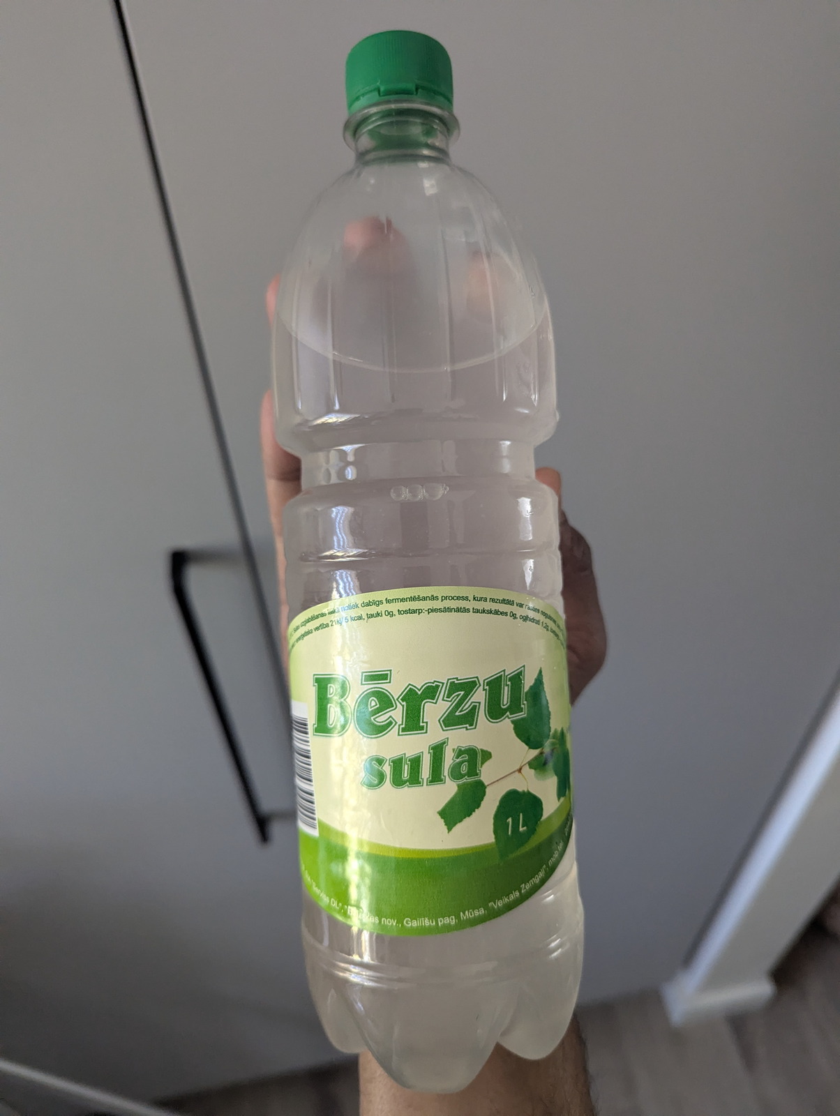 1 litro PET butelis Bērzu sulos (latviškos beržų sulos) su žaliomis lapelių piešiniais etiketėje, laikomas prie virtuvės sienos